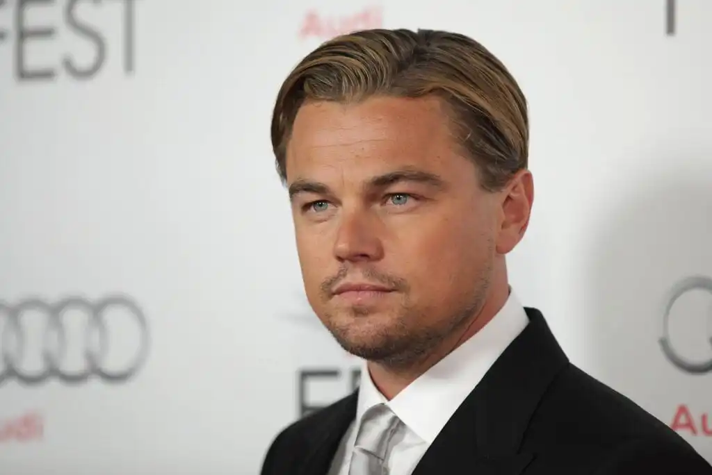 A man in a black suit, white shirt, and silver tie poses against a white backdrop with blurred logos. He has slicked-back dark blond hair and a trimmed beard, and he is looking slightly to the side.