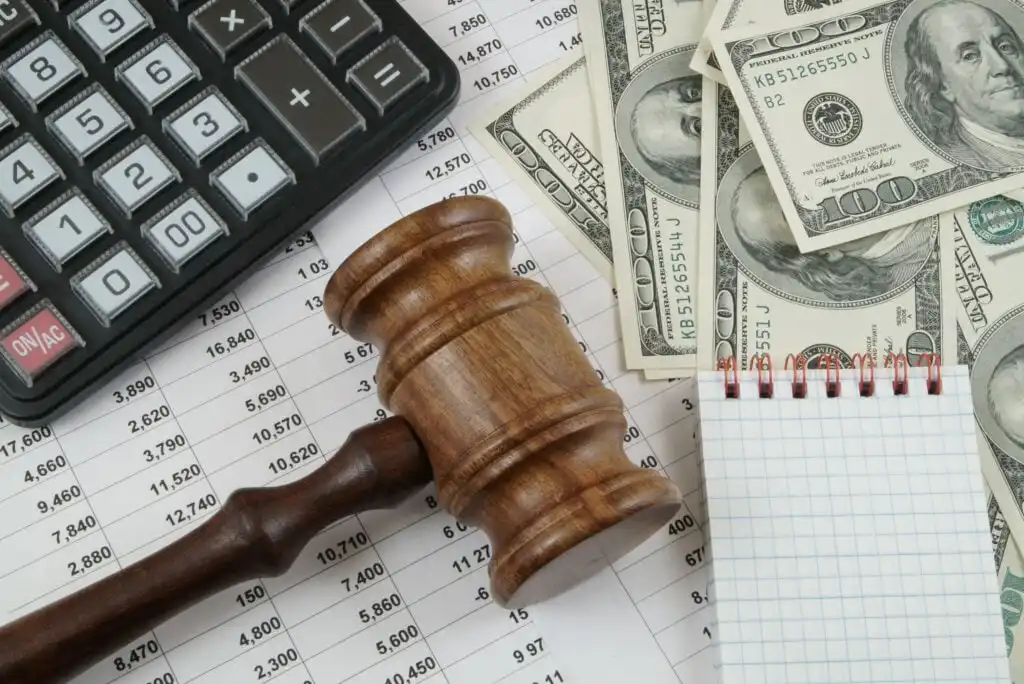 A wooden judge’s gavel, calculator, U.S. dollar bills, and a blank notepad rest on financial spreadsheets with numbers. The image suggests themes of finance, law, or legal decisions involving money.