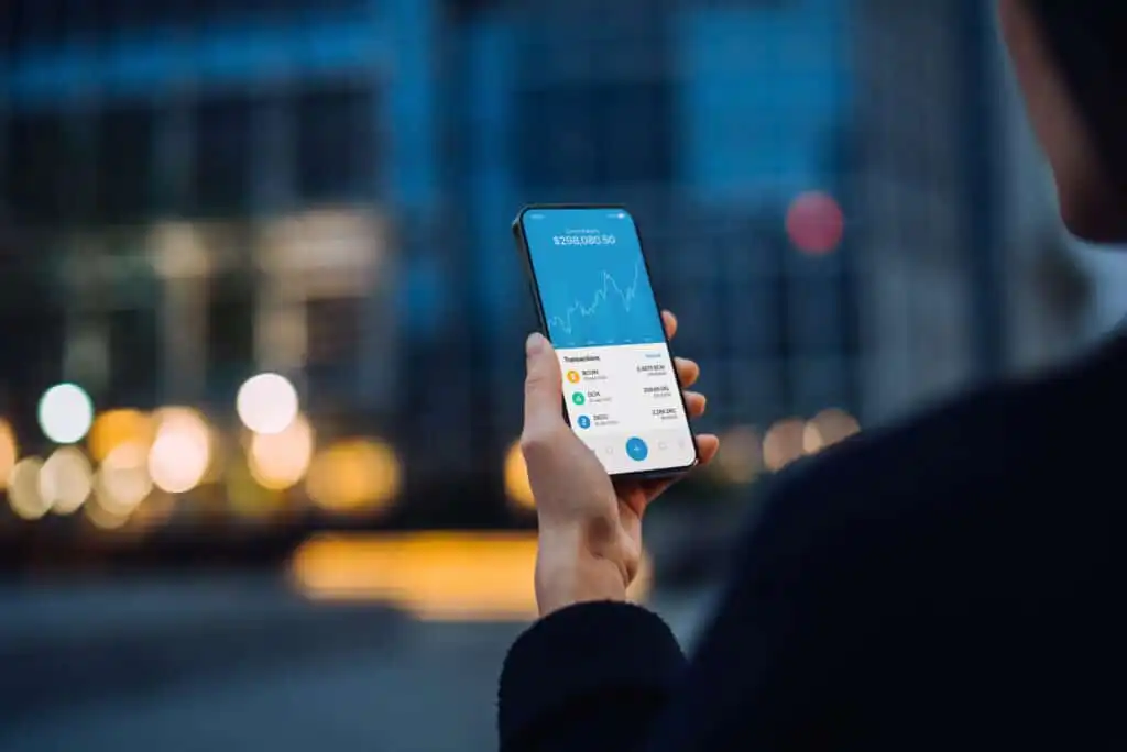 A person holds a smartphone displaying a cryptocurrency investment app with a chart and various coin balances, against a blurred urban background with lights.