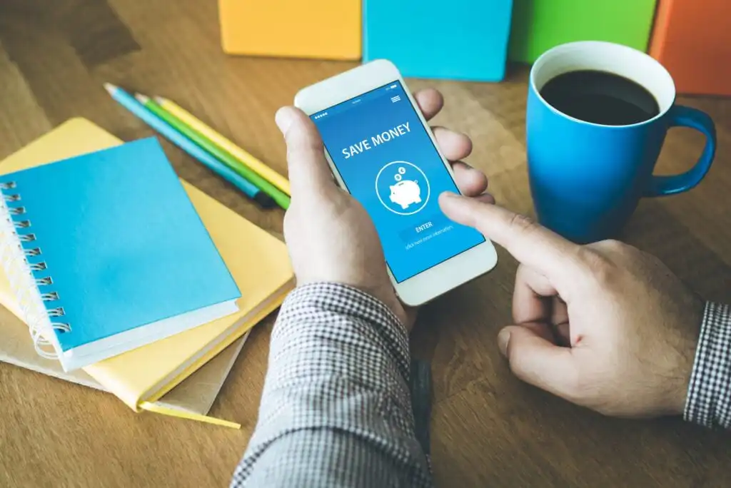 A person uses a smartphone app with a “Save Money” screen showing a piggy bank icon. Nearby are a blue notebook, yellow and green pencils, and a cup of coffee on a wooden desk.