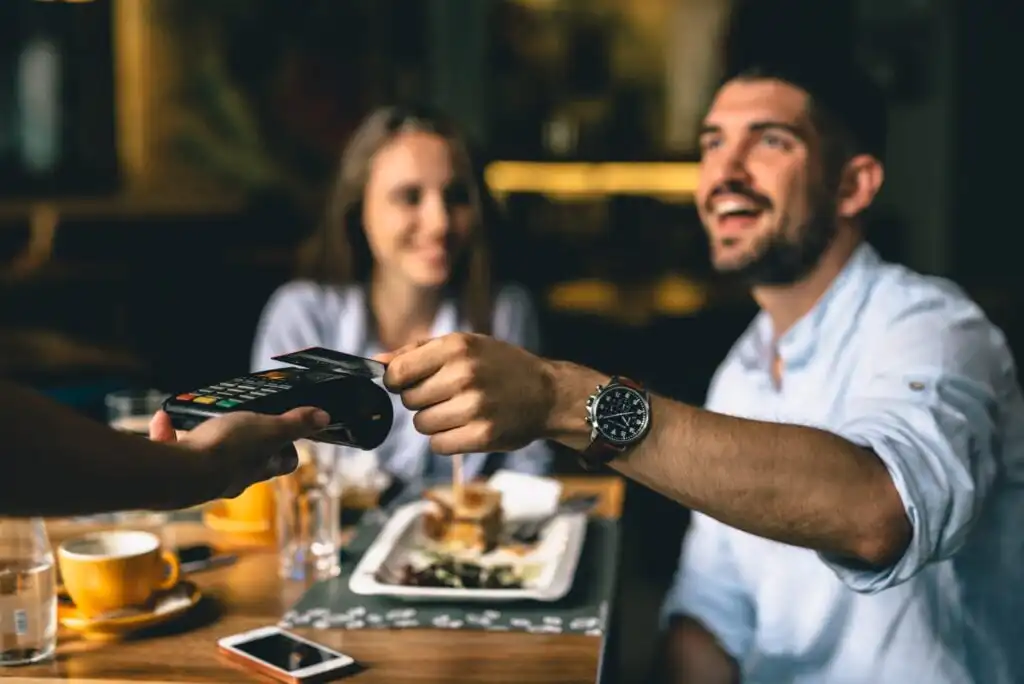 A man smiles as he pays with a credit card using a contactless payment terminal at a restaurant table, while a woman sitting across from him also smiles. Food and drinks are visible on the table.