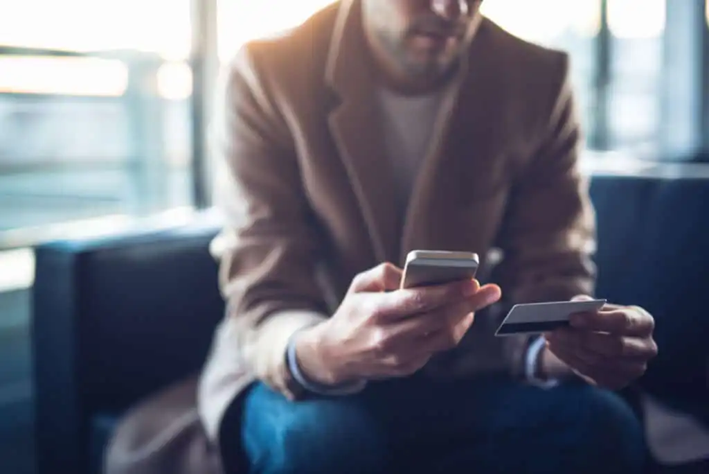A man in a brown coat sits indoors, holding a smartphone in one hand and a credit card in the other, appearing to make an online transaction or payment.