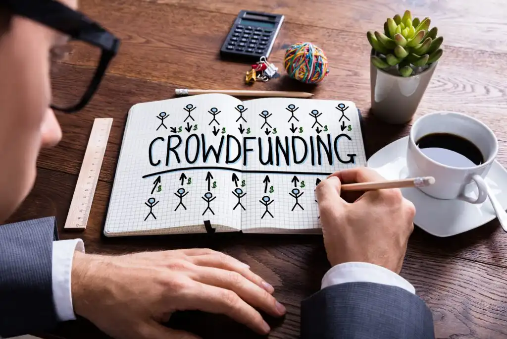 A person in a suit writes CROWDFUNDING in a notebook with stick figure drawings around the word. Nearby are a coffee cup, plant, calculator, rubber band ball, and ruler on a wooden desk.