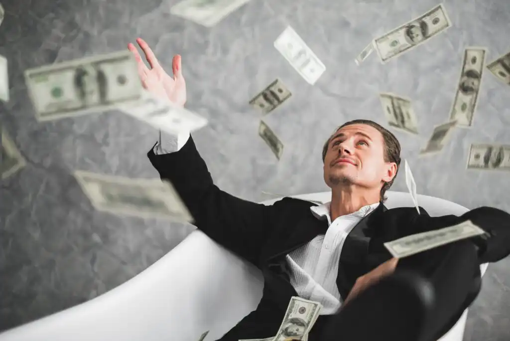 A man in a suit lounges in a white bathtub while tossing and surrounded by falling U.S. dollar bills, looking upward with a pleased expression.