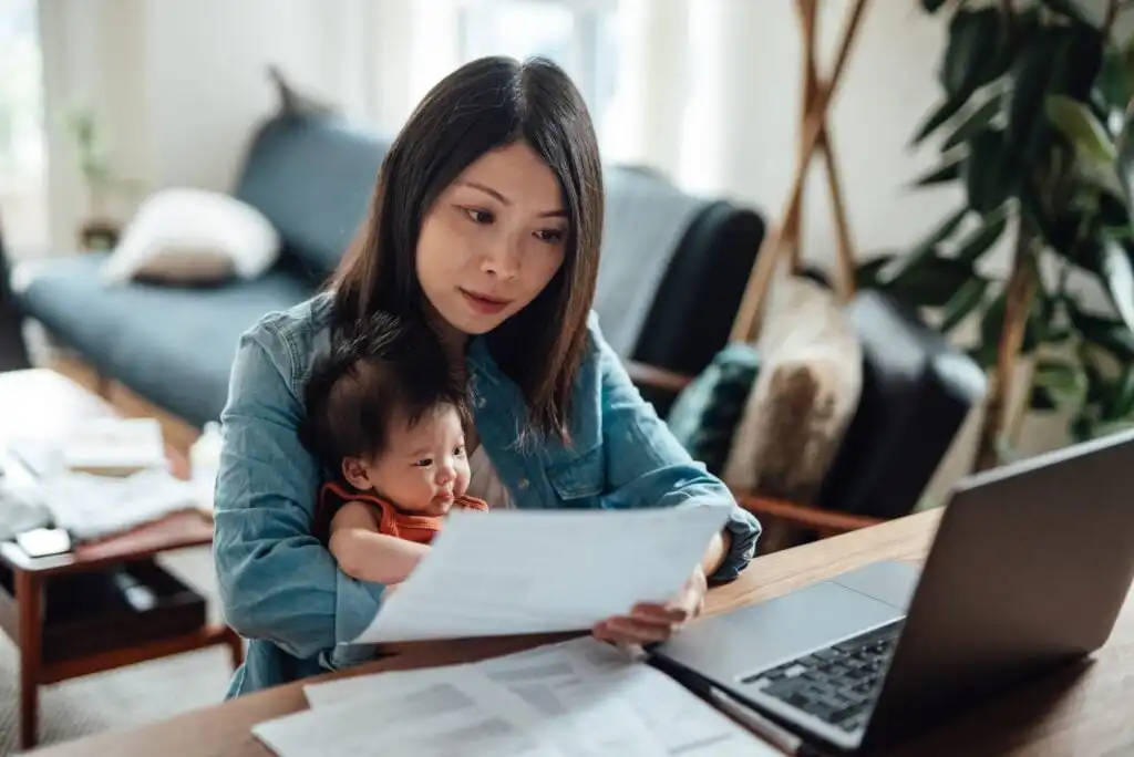 A woman holds a baby in her lap while looking at papers and working on a laptop at a table in a living room, appearing focused and busy.