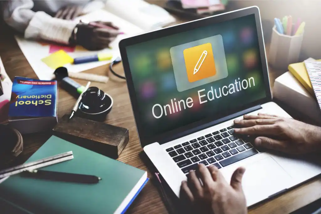 A person uses a laptop displaying the words Online Education on the screen, surrounded by notebooks, colored folders, a camera lens, and stationery on a cluttered desk.