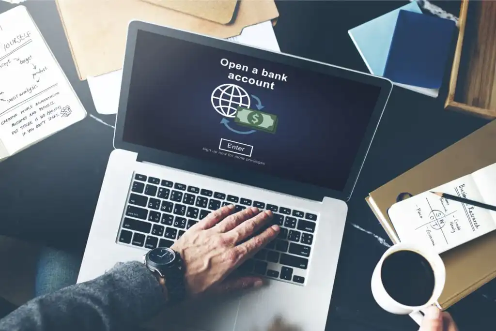 A person’s hand on a laptop keyboard displaying a screen that says “Open a bank account” with icons of a globe and dollar bill. The desk has papers, notebooks, a phone, and a cup of coffee.