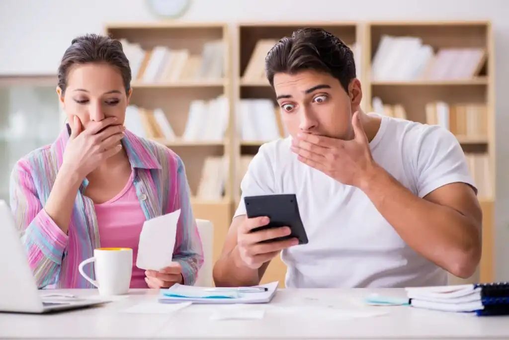 A surprised man holds a smartphone and covers his mouth, while a woman beside him covers her mouth and looks at a paper. They sit at a table with documents, a laptop, and a cup in a room with shelves in the background.