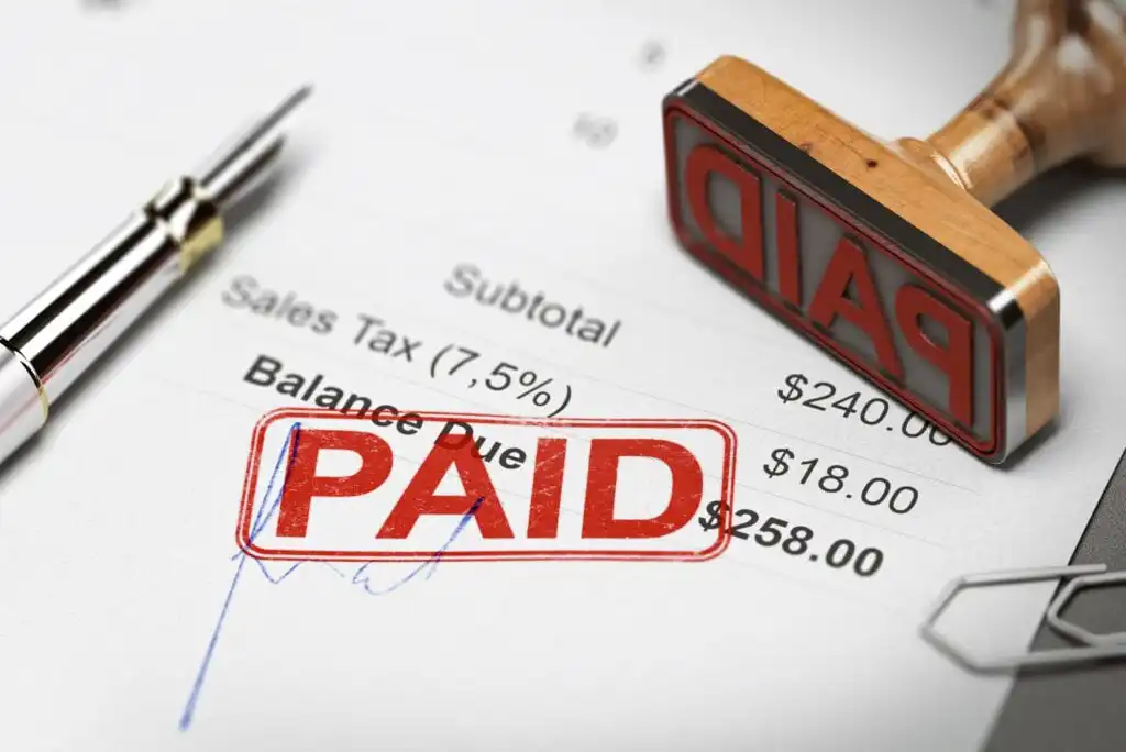 A close-up of an invoice stamped PAID in red ink, with a pen and a rubber stamp nearby. The invoice shows sales tax, subtotal, and a zero balance due.