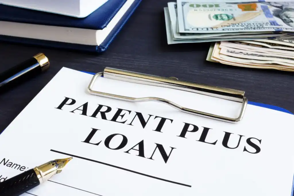 A clipboard holds a document labeled Parent PLUS Loan next to a pen, stacked notebooks, and a pile of U.S. cash on a dark desk.