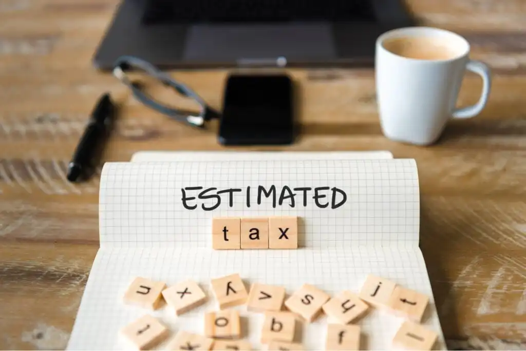 A notebook with ESTIMATED written on it, and scrabble tiles spelling tax on the page. Nearby are a pen, eyeglasses, smartphone, laptop, and a cup of coffee on a wooden desk.