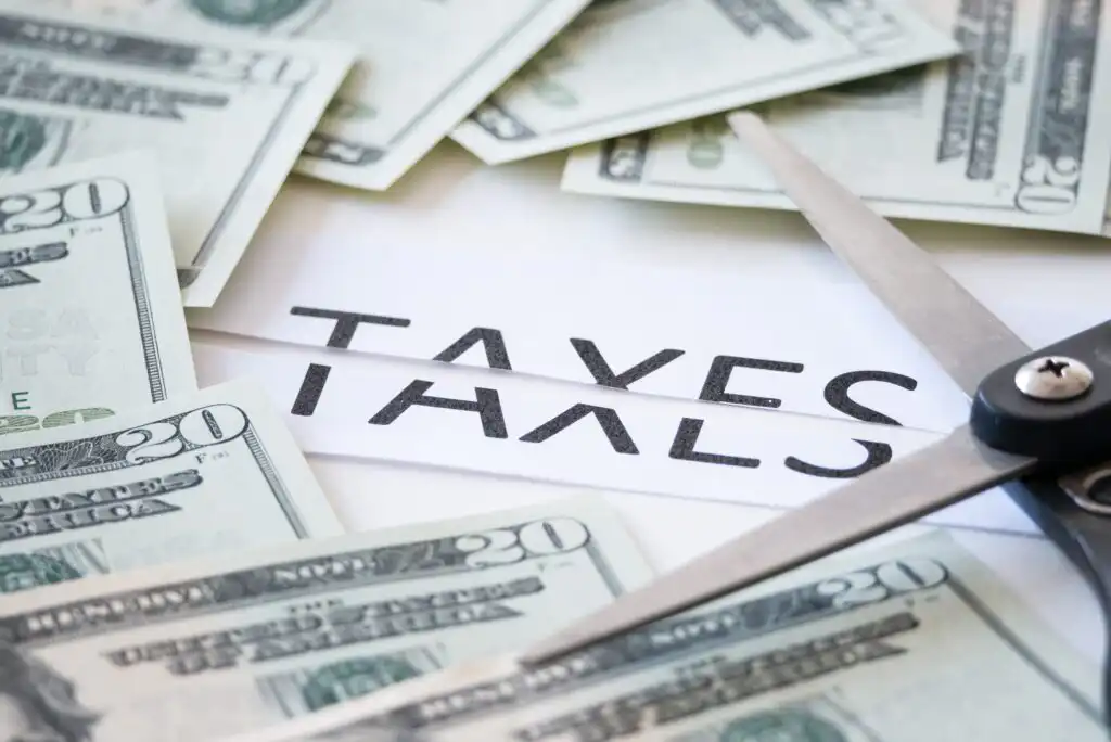A pair of scissors cutting through a paper labeled TAXES, surrounded by spread out twenty-dollar bills, symbolizing tax reduction or tax cuts.