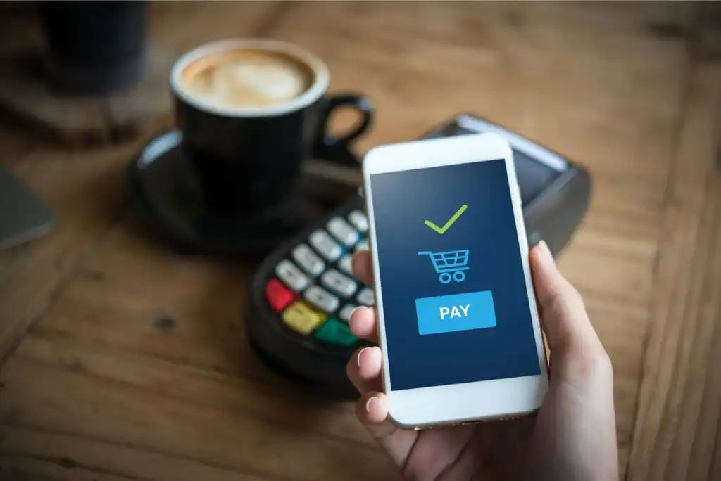 A person holds a smartphone displaying a payment confirmation screen with a shopping cart icon and PAY button, near a card payment terminal on a wooden table with a cup of coffee in the background.