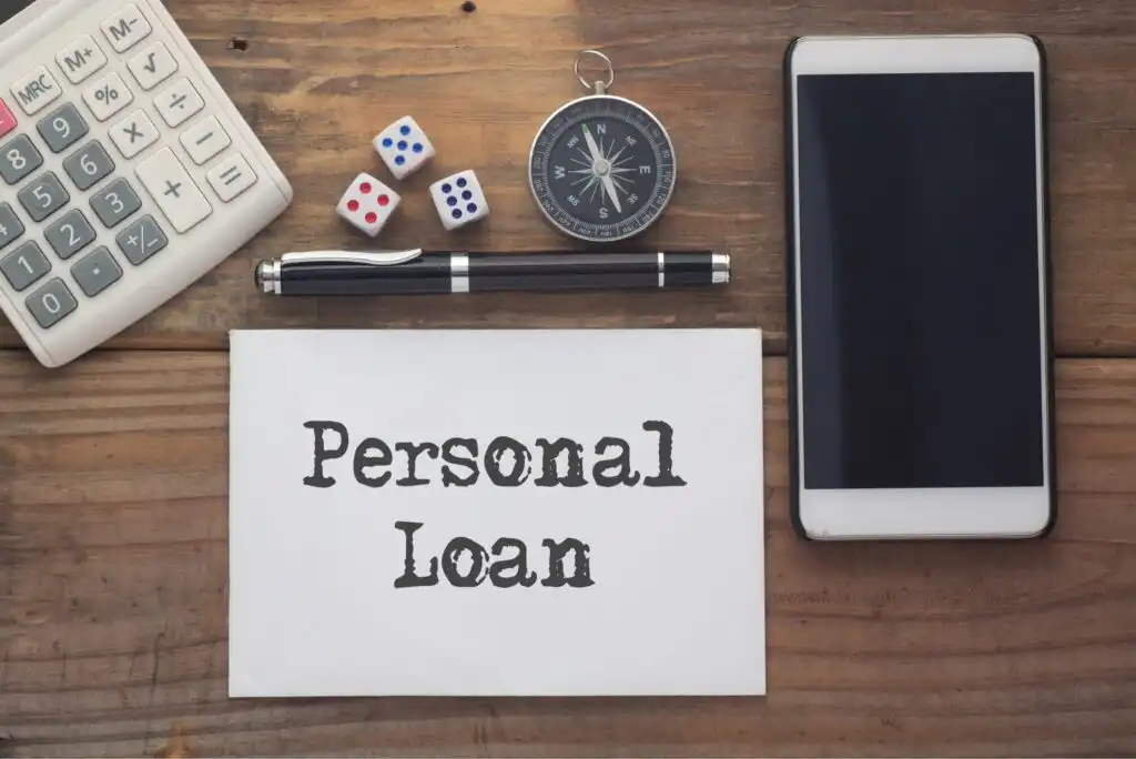 A paper with Personal Loan written on it is on a wooden desk, surrounded by a calculator, pen, three dice, a compass, and a smartphone.