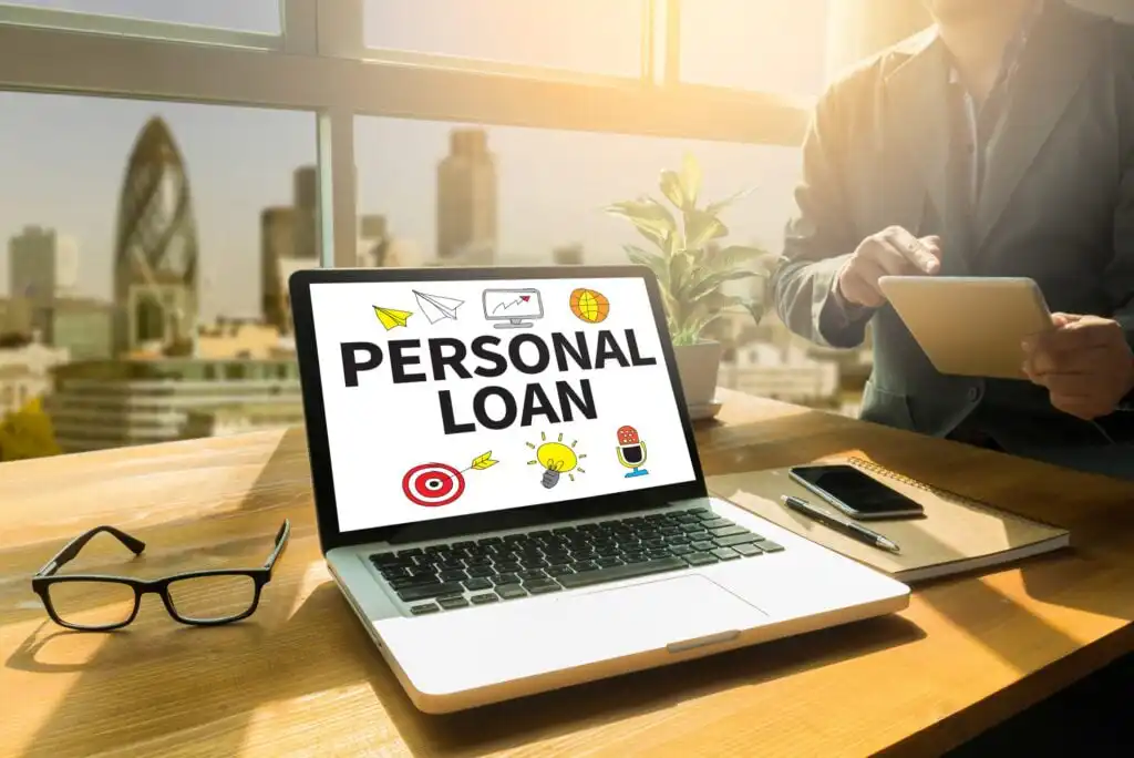 A laptop on a desk displays the words PERSONAL LOAN on its screen. Nearby are glasses, a notebook, pen, and a person holding a tablet. A city skyline is visible through large windows in the background.