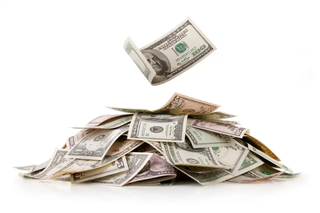 A pile of assorted U.S. dollar bills sits on a white surface, with one bill floating in the air above the pile.