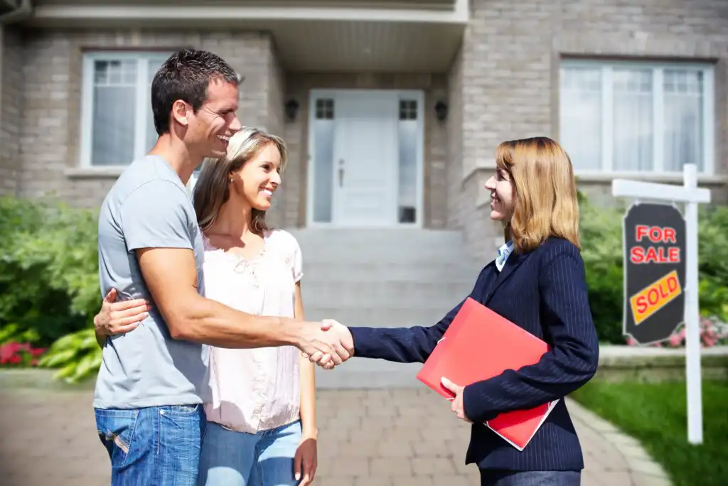 A smiling couple stands outside a house, shaking hands with a real estate agent holding a red folder; a “For Sale” sign with a “Sold” sticker is visible in the yard.