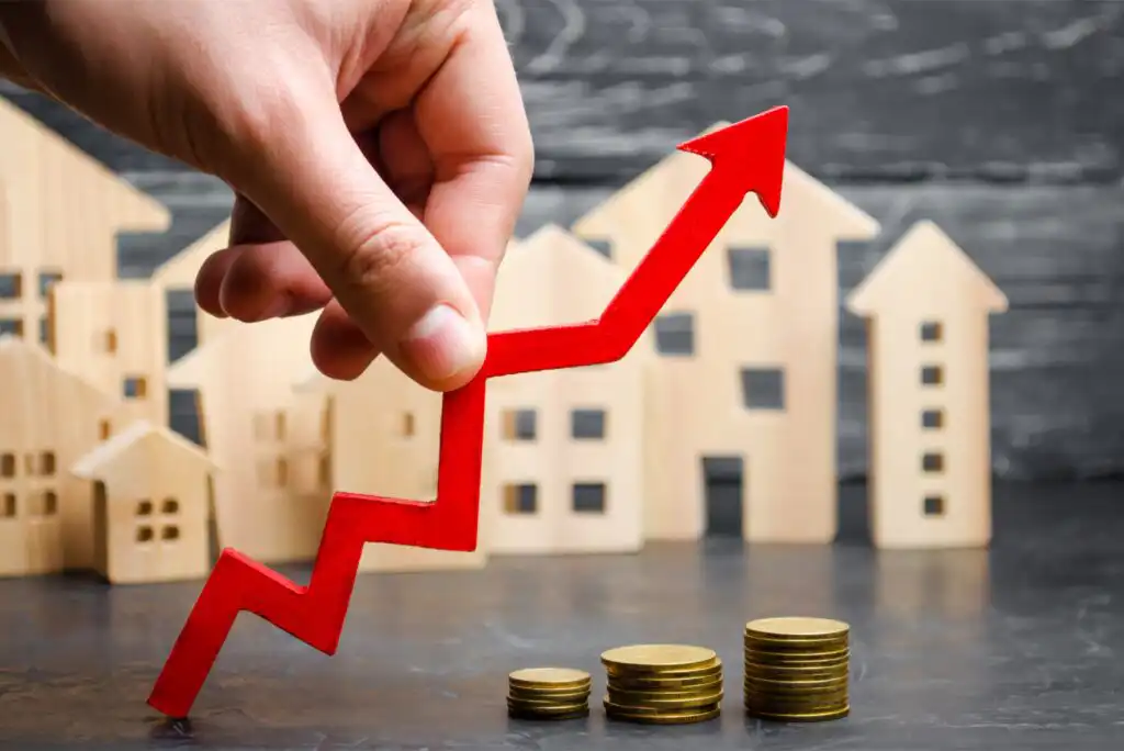 A hand holds a red upward arrow in front of wooden house models and stacks of coins, symbolizing rising real estate prices or property investment growth.