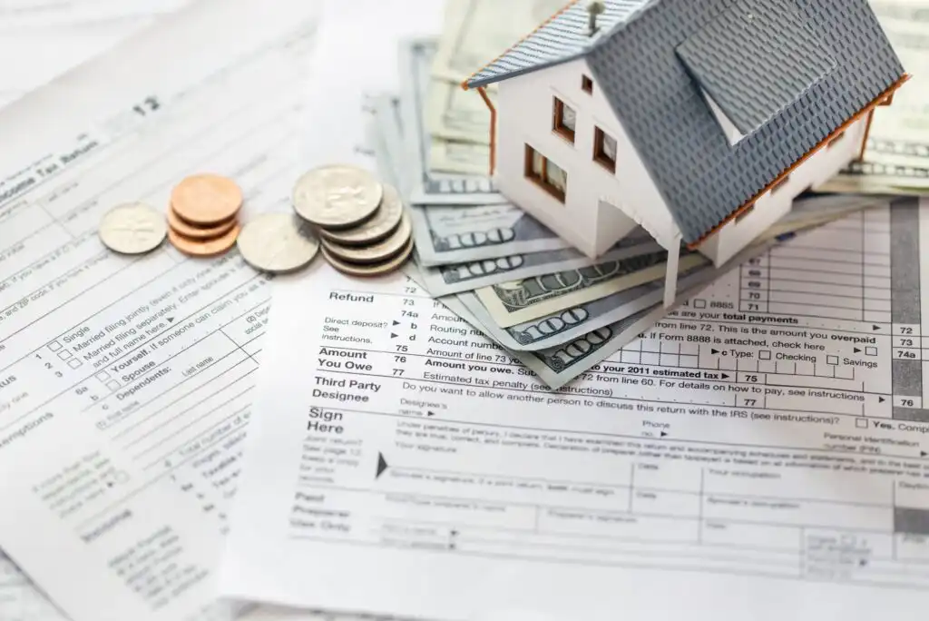 A small model house sits on top of a stack of dollar bills, coins, and tax forms, symbolizing real estate, taxes, and financial planning.