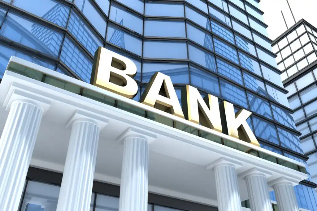 A modern bank building with glass windows and white columns, featuring a large yellow BANK sign above the entrance.