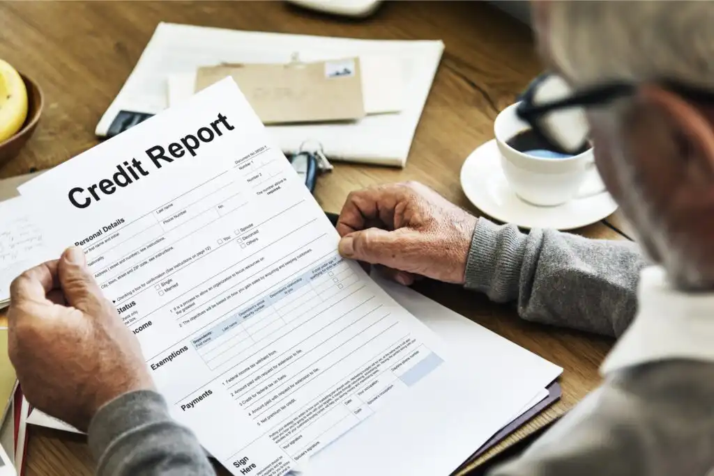 A person holding and reading a credit report document at a desk, with paperwork, a pen, a white coffee cup, and an envelope in the background.