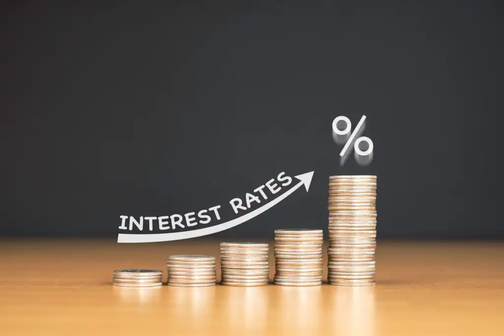 Stacks of coins arranged in increasing height, with an upward arrow labeled Interest Rates and a large percent sign, symbolizing rising interest rates against a dark background.