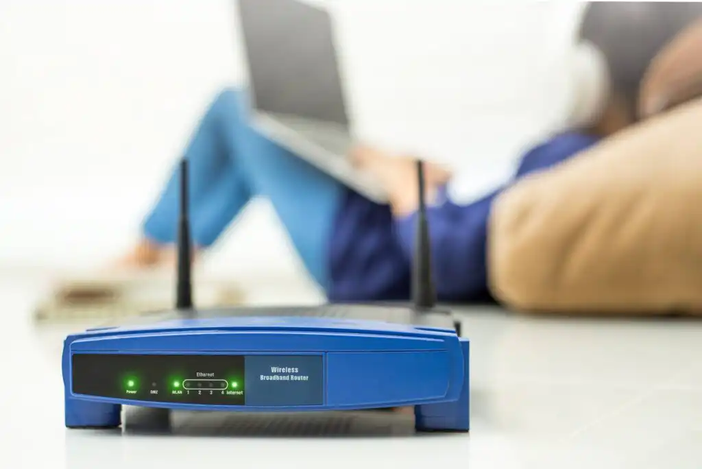 A blue wireless router with indicator lights is in focus on a white surface. In the blurred background, a person sits on the floor using a laptop, wearing headphones and leaning on a cushion.