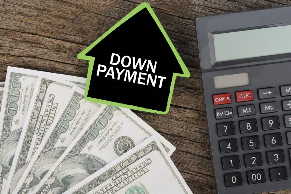 A calculator, several hundred-dollar bills, and a black house-shaped sign with a green border and the words DOWN PAYMENT are arranged on a wooden surface.