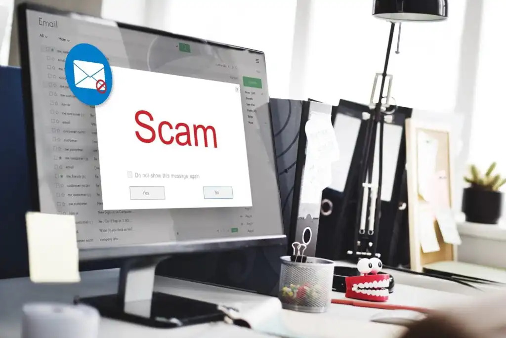 A computer screen displays a warning message with the word Scam in large red letters and email inbox visible in the background, indicating a phishing or scam alert.