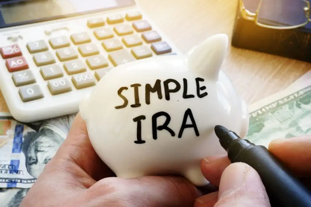 A person writes SIMPLE IRA on a white piggy bank with a black marker. A calculator, cash, and documents are visible on a wooden desk in the background.