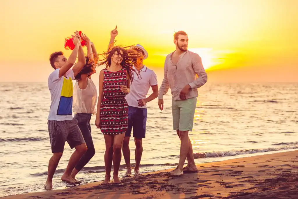 Five young adults enjoy a sunset on the beach, smiling and dancing together near the shoreline, with the sun setting over the calm sea in the background.