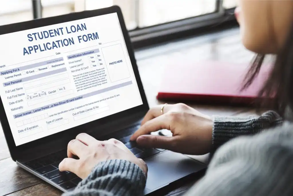 A person types on a laptop displaying an online student loan application form. A closed red notebook and a window are visible in the background.