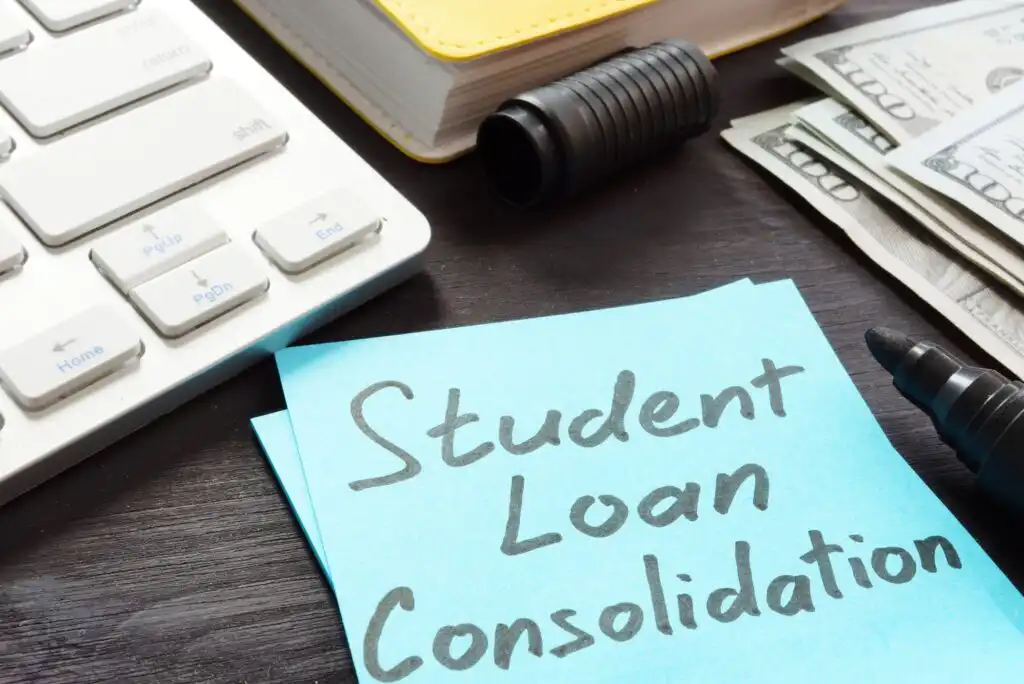 A desk with a sticky note reading Student Loan Consolidation, a keyboard, a black marker, a stack of cash, and a closed notebook, suggesting financial planning and loan management.