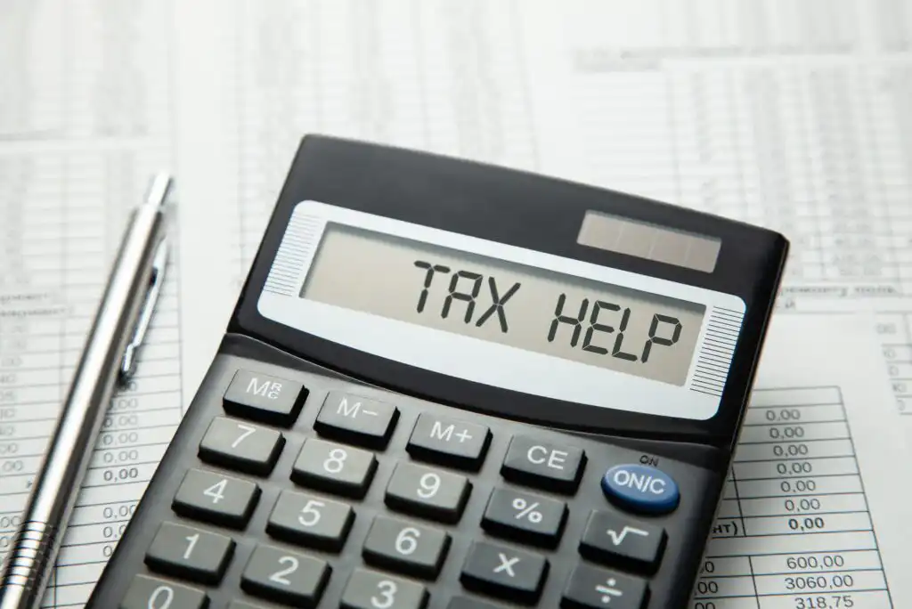 A calculator with the words TAX HELP displayed on its screen sits on top of financial documents, next to a silver pen.