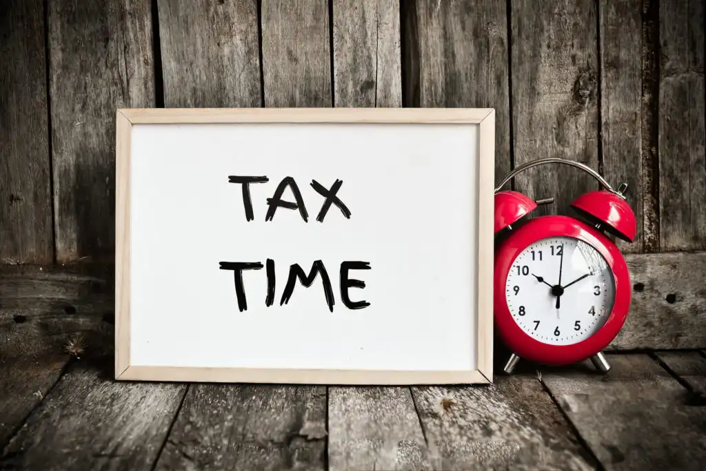 A whiteboard with the words TAX TIME next to a red vintage alarm clock showing 10:10, set on a wooden surface with a rustic wooden background.