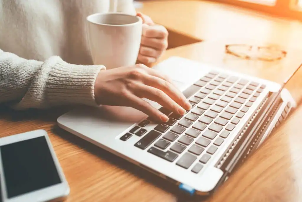 A person wearing a light sweater holds a white mug in one hand while typing on a laptop with the other. A smartphone and eyeglasses rest on the wooden desk beside them.