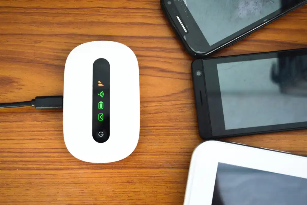 A white portable Wi-Fi hotspot device with indicator lights is charging on a wooden surface, surrounded by three smartphones and a tablet with black screens.
