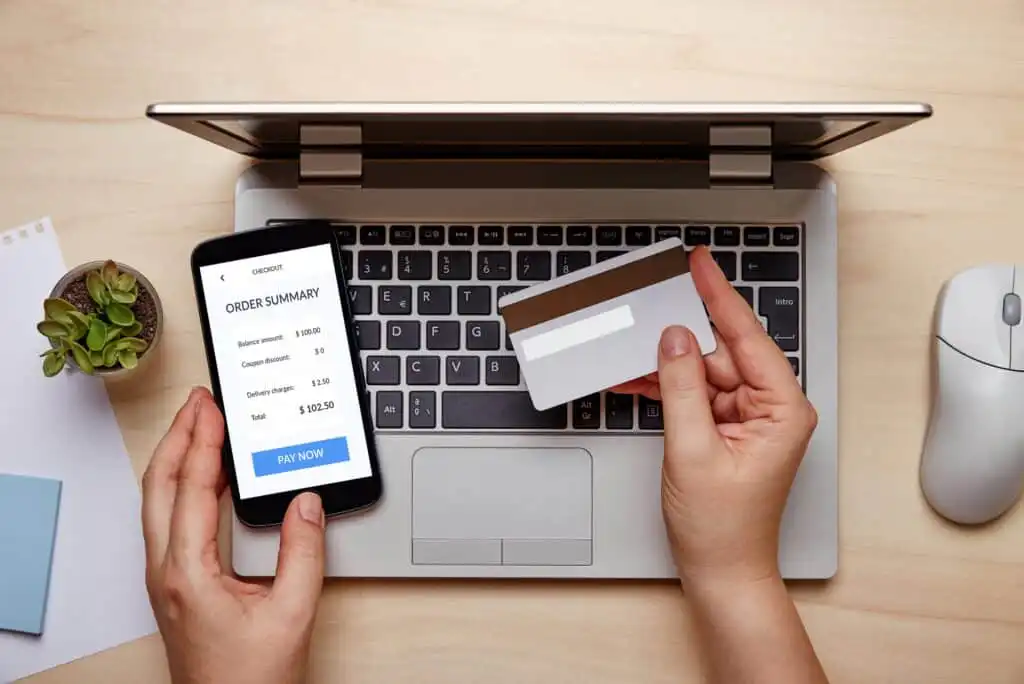 A person holding a credit card in one hand and a smartphone displaying an online order summary in the other, sitting in front of a laptop on a desk with a plant and a computer mouse nearby.