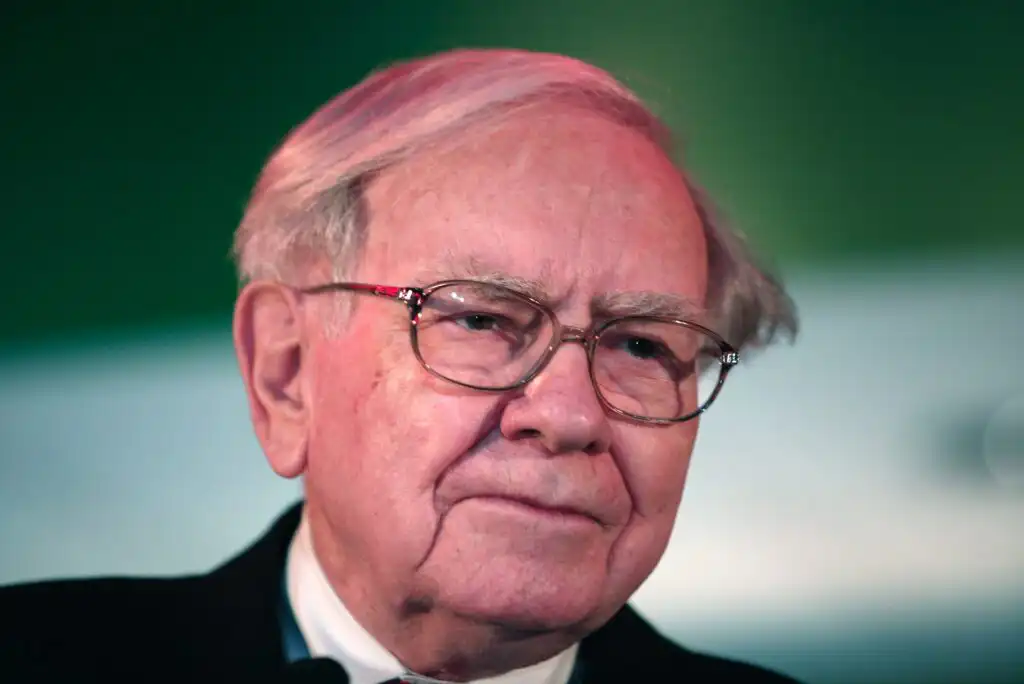 An elderly man with white hair and glasses wearing a suit and tie, looking thoughtful. The background is blurred in shades of green and white.