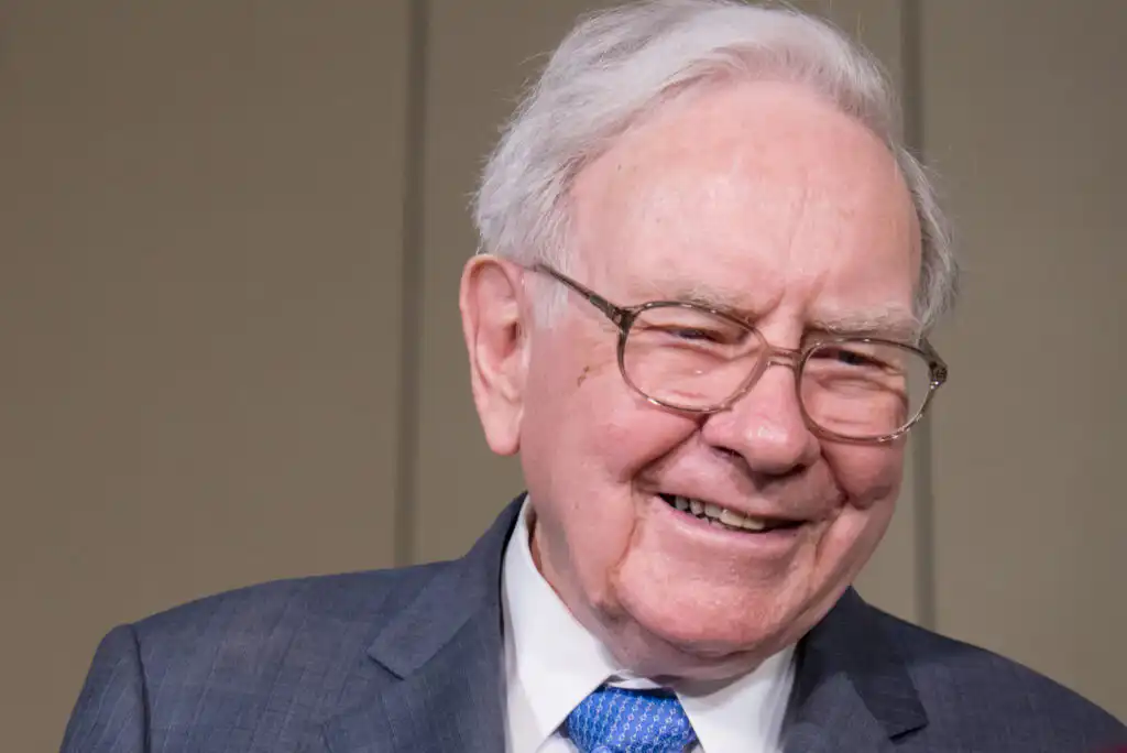 An elderly man with white hair and glasses, wearing a blue suit, white shirt, and blue patterned tie, smiles warmly against a plain background.