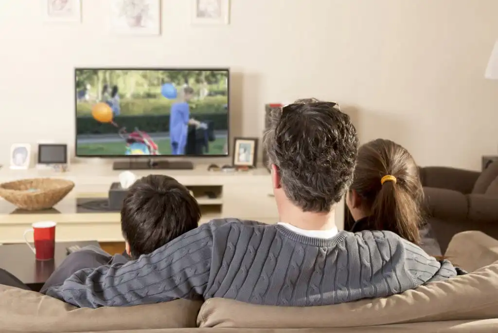Three people sit on a couch watching TV together in a cozy living room. The TV displays an outdoor scene, and there are cups and decorative items on the table in front of them.