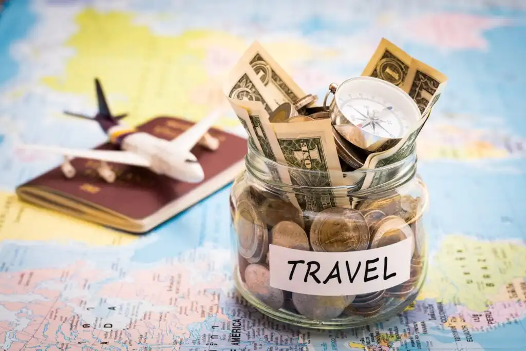 A glass jar labeled Travel filled with coins and dollar bills, next to a compass. In the background is a map, a toy airplane, and a passport.