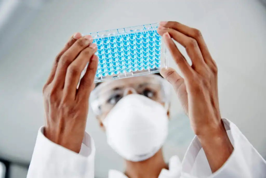 A scientist wearing a mask, safety goggles, and lab coat holds up a clear plastic plate filled with blue samples, examining it closely in a laboratory setting.