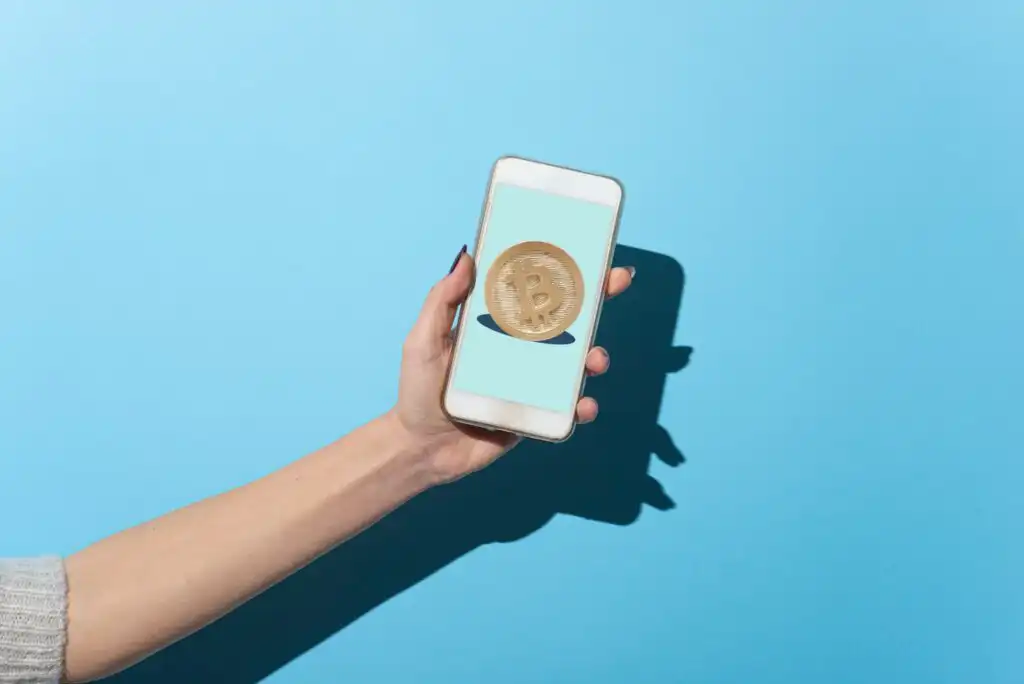 A hand holding a smartphone against a blue background, with a large gold Bitcoin symbol displayed on the phone screen.