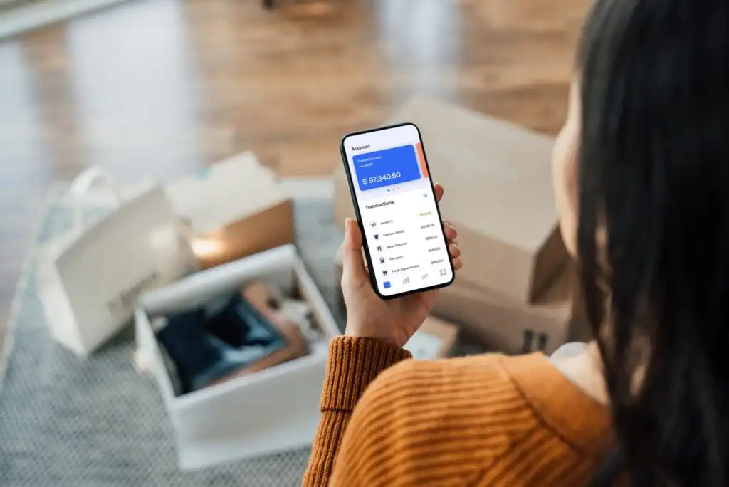 A person in an orange sweater checks their digital wallet balance and transactions on a smartphone, sitting near shopping bags and an open box on a carpeted floor.