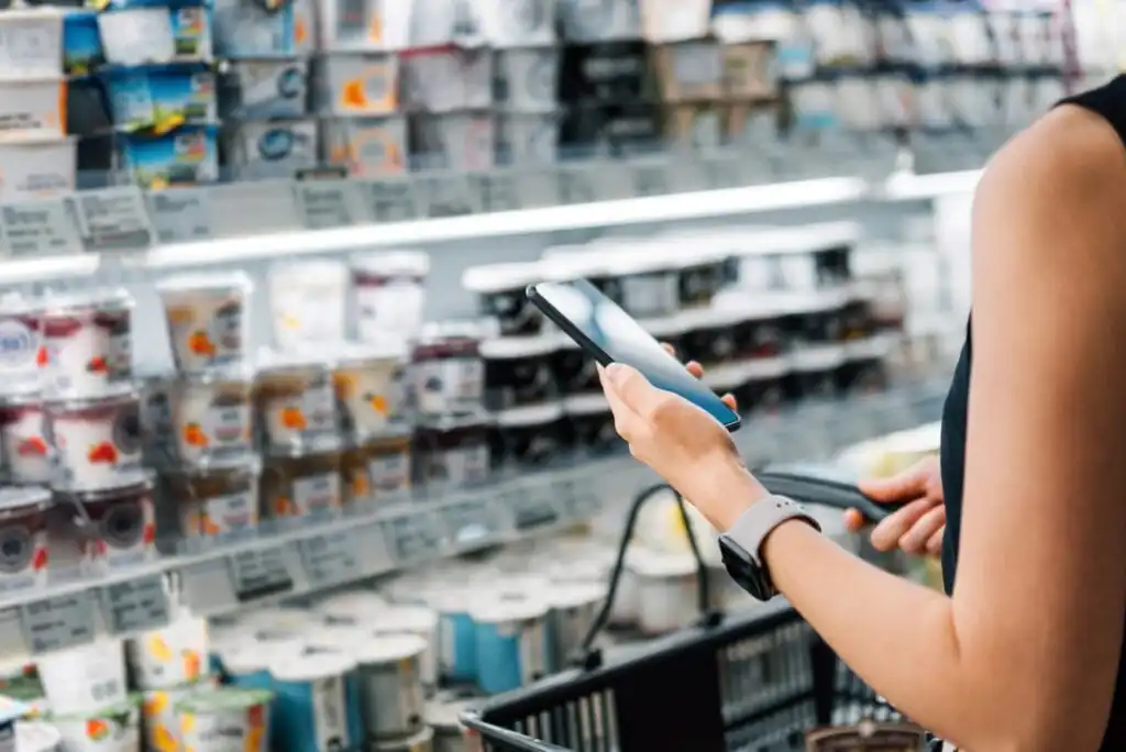 A person holding a smartphone and shopping cart stands in front of a refrigerated grocery shelf stocked with various yogurt containers, possibly scanning or comparing products.