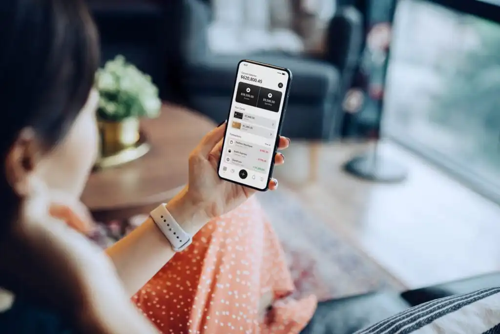 A person sitting indoors, wearing a smartwatch and holding a smartphone. The phone screen displays a financial app with account balances and transaction details. The background shows a blurred living room setting.