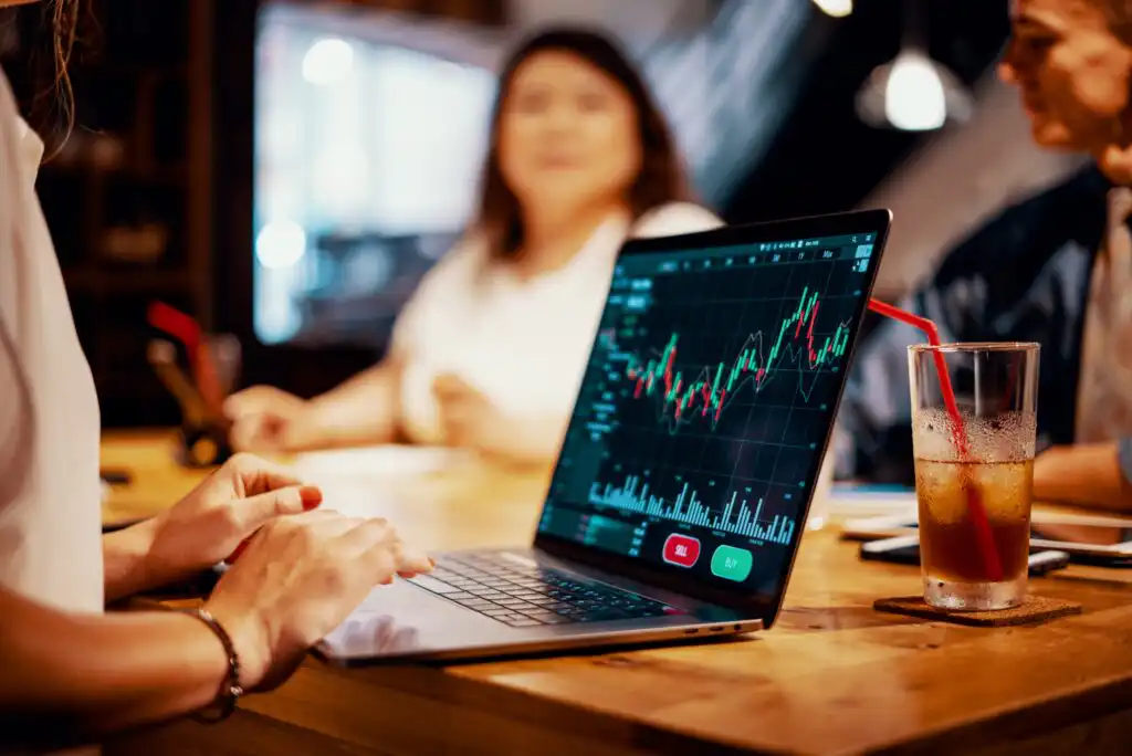 A person uses a laptop displaying a colorful stock market chart in a meeting setting, with two blurred people and a drink with a straw in the background.