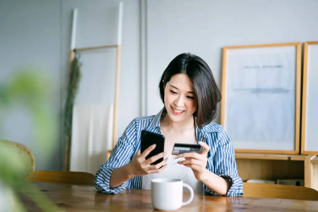 A woman sits at a table smiling, holding a smartphone in one hand and a credit card in the other, with a white mug in front of her in a bright, modern room.
