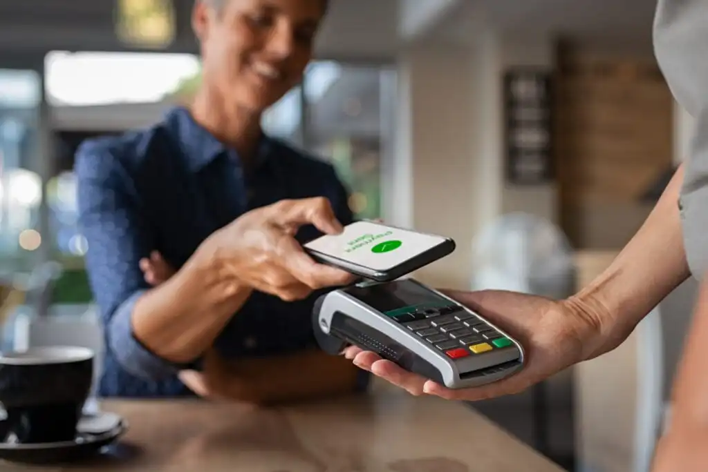 A person smiles while making a contactless payment with a smartphone at a card reader, held by another person, in a cafe setting. A coffee cup is visible on the table.